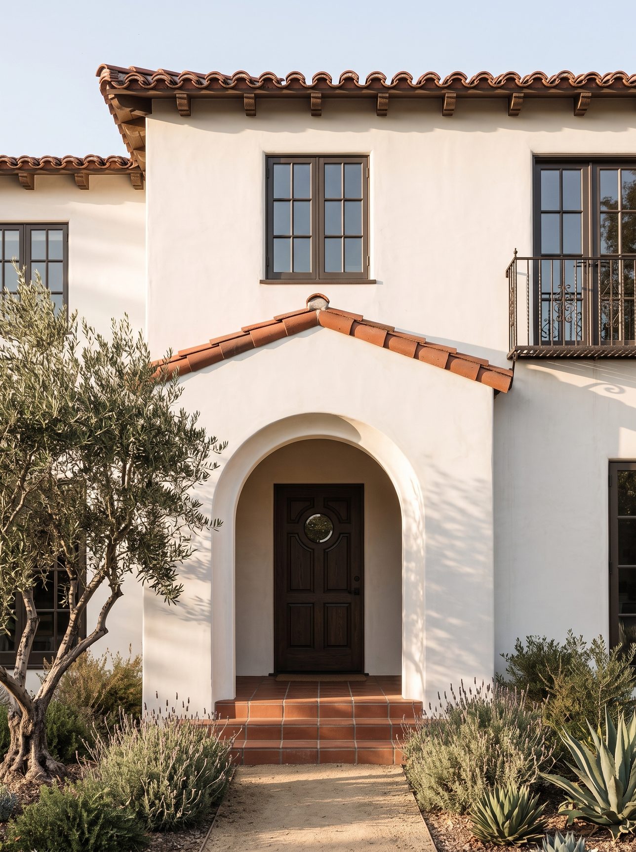 The front of the Reeves house: a white two-story Spanish Revival with a clay tile roof, its dark wooden door set back in an arched porch behind an olive tree and dry-garden planting