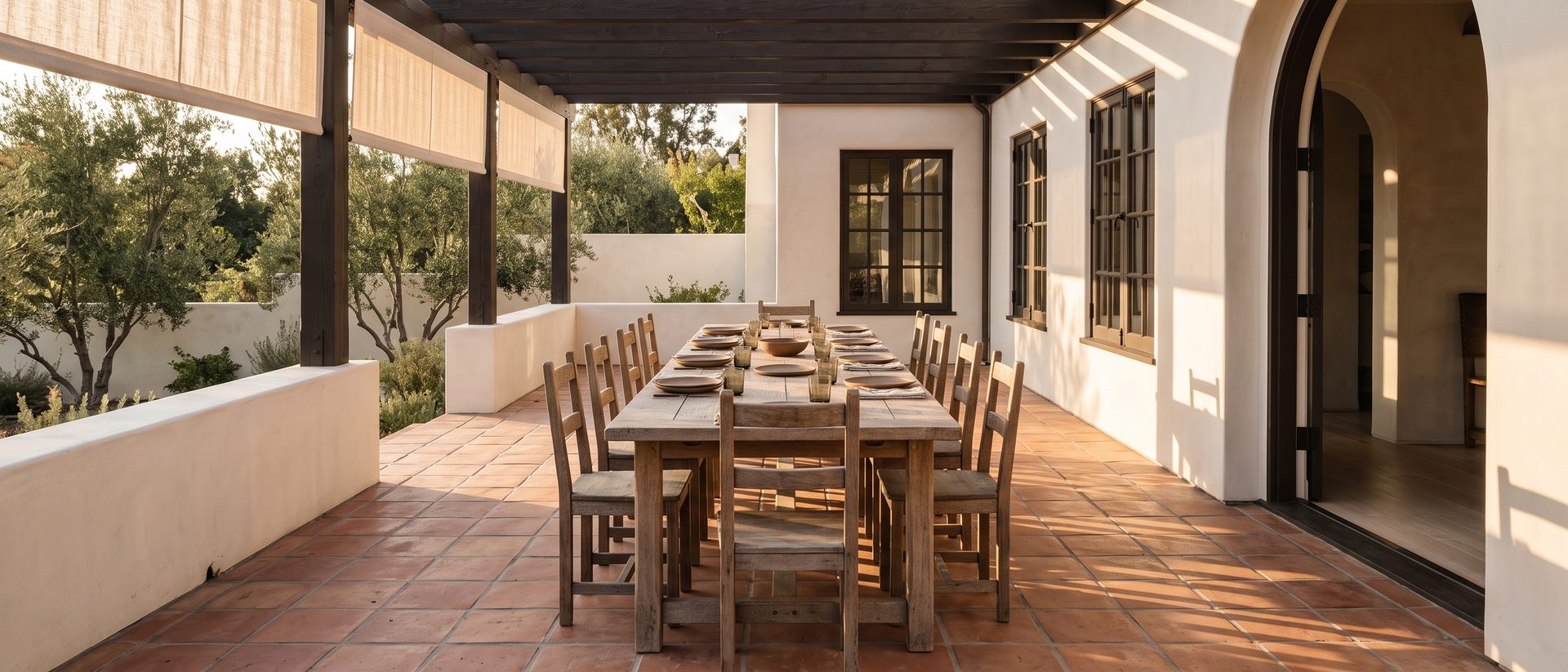 The terrace: a long weathered oak table set for a crowd on terracotta tile, under a timber pergola hung with linen shades