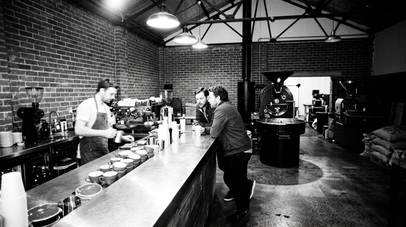 Inside the shop: a barista behind a long steel bar lined with cups, two customers leaning on it, and the black drum roaster beyond, under bare pendant lamps and brick walls