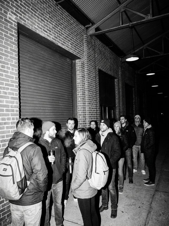A line of people in coats and beanies waiting along the brick front before the roll-up door opens, a few holding travel mugs, their breath showing in the cold