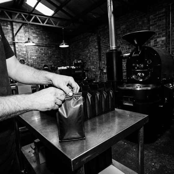 Hands folding the top of a plain black coffee bag on a steel table, a row of filled bags beside it and the roaster behind