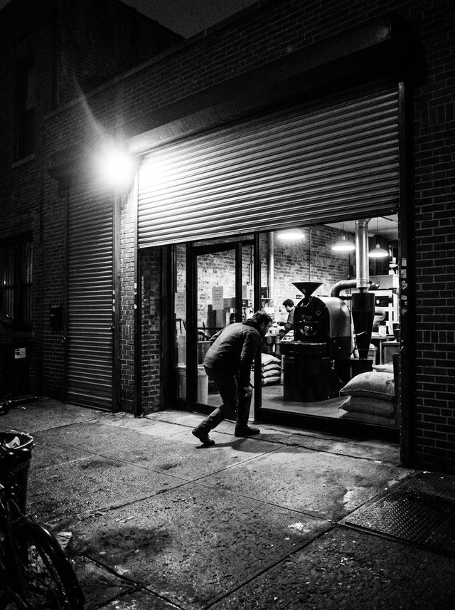 The shop at night: the roll-up door half raised and lit from inside, a customer ducking under it past the roaster and stacked sacks of green coffee