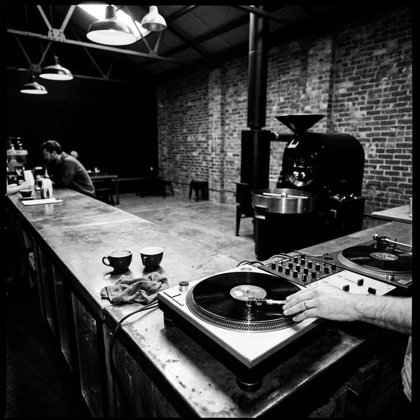 A hand setting the needle on a record at the end of the steel bar, two espresso cups beside the turntable and the roaster across the room